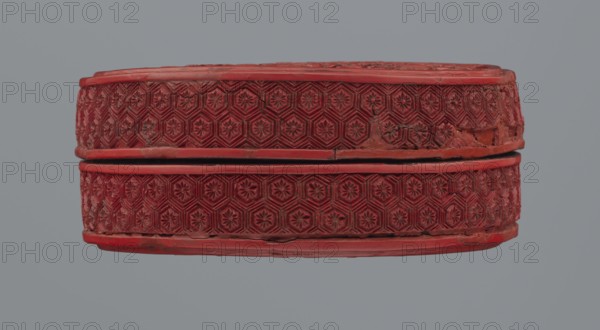 Box with lid, Chinese; Qing dynasty 1644–1911; two-tone lacquer red and black lacquer inside; 19th century; circular container decorated with floral ornament—peonies, bamboos, peaches—and kikko geometric pattern; base: height 2.1 cm, diameter 8.8 cm; lid: height 1.8 cm, diameter 8.8 cm; box height 3.8 cm; function: container., A red oval box with an ornate repeating geometric floral pattern carved around its sides, showing a removable lid separated by a narrow dark gap; the carved surface has worn areas and irregularities, with visible rough edges and small chips. The background is a uniform gray. The visible colors are red and gray with some darker red and blackish shadows in the carved recesses.