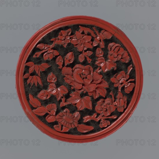 Box with lid;, Two-tone lacquer red and black lacquer inside. 19th century, Qing 1644–1911, China. Container/box with floral ornament peonies, bamboos, peaches, kikko geometric pattern. Dimensions: base: height 2.1 cm, diameter 8.8 cm; lid: height 1.8 cm, diameter 8.8 cm; box height 3.8 cm; diameter 8.9 cm., A round carved object with a raised rim in red surrounding an inner carved floral pattern in red against a dark textured background, set on a grey surface; the carving shows a group of overlapping flowers and leaves with detailed petals and veins, and the overall visible colors are red, dark brown or black, and grey.
