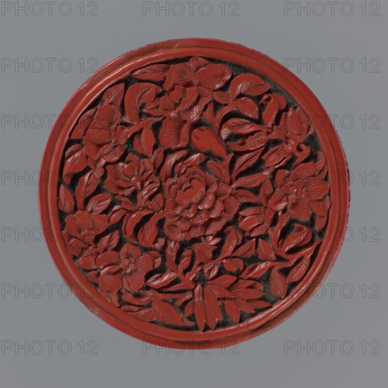 Box with lid, Chinese, Qing dynasty 1644–1911, 19th century. Two-tone lacquer red and black; black lacquer inside. Decorated with floral ornament: peonies, bamboos, peaches iconography, kikko geometric pattern. Function: container. Dimensions: base height 2.1 cm, diameter 8.8 cm; lid height 1.8 cm, diameter 8.8 cm; overall box height 3.8 cm, diameter 8.9 cm., A round carved object with a raised floral relief occupying the center, featuring a group of intricately carved flowers and many leaves arranged densely; the carved elements are predominantly red with darker recesses between petals and leaves creating contrast, and the piece is framed by a red circular rim set against a gray background.