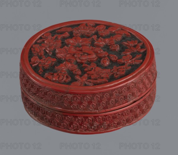 Box with lid; Chinese; Two-tone lacquer red and black lacquer inside; floral ornament with peonies, bamboos, peaches; kikko geometric pattern; Qing dynasty 1644–1911; 19th century; maker, container. Dimensions: base height 2.1 cm, diameter 8.8 cm; lid height 1.8 cm, diameter 8.8 cm; box height 3.8 cm, diameter 8.9 cm., A round carved object with a raised lid featuring a dense floral relief, showing a group of stylized flowers and leaves in a glossy red over a darker background; the sides have repeated geometric carved patterns wrapping around the circumference, also in red with darker recesses; the overall colors visible are red, dark gray or black in the carved recesses, and a neutral gray background behind the object.