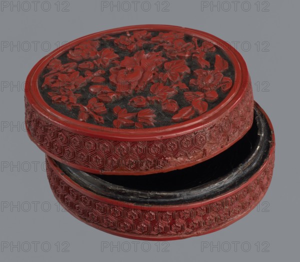 Box with lid, Chinese, Qing dynasty 1644–1911, 19th century. Two-tone lacquer red exterior, black lacquer interior. Decorated with floral ornament—peonies, bamboos, peaches—and kikko geometric pattern. Container box: height 3.8 cm, diameter ~8.8–8.9 cm. Lid: height 1.8 cm, diameter 8.8 cm. Base: height 2.1 cm, diameter 8.8 cm., Function: container., A round carved box with a slightly open lid showing a dark interior, the box and lid covered in raised floral and geometric carvings; the dominant color is red with visible black in the recessed areas and inside the box, and a grey background behind the box.