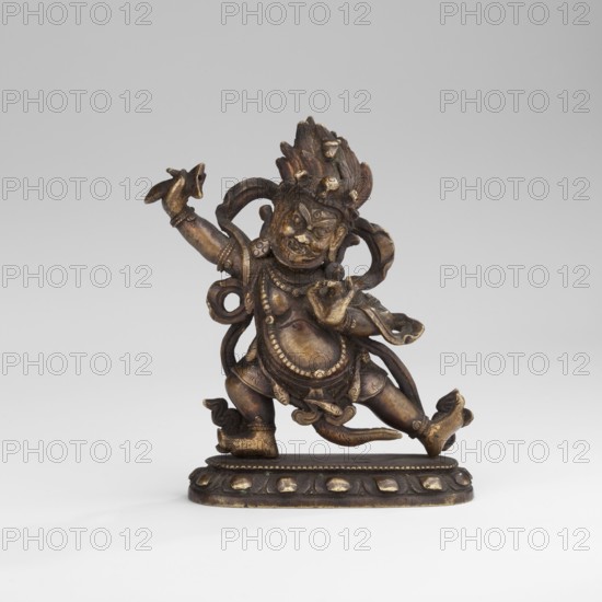 Hayagriva, unknown sculptor, Buddhist art, Himalayan art, Tibetan Buddhist art, Himalayan culture, dharmapala iconography, 2nd half of the 18th century, Tibet, sculpture, figurine; height 7.0 cm, width 6.1 cm, depth 2.4 cm, A small bronze figure with a dark brown and golden patina stands on an oval base; the figure has a dynamic, dancing pose with one leg raised and one arm extended upward, wearing ornate jewelry and draped cloth elements, and a headdress with textured, flame-like details. The surface shows areas of worn gold highlighting on raised features, with darker brown recesses throughout. The base is dark brown with golden accents on rounded decorative elements, and the background is a smooth light gray.