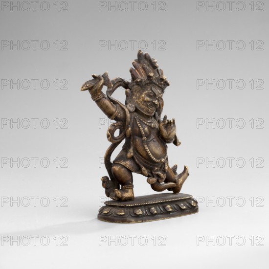 Hayagriva, unknown sculptor, 2nd half of the 18th century, Tibet; Buddhist, Tibetan/Himalayan art; dharmapala iconography; sculpture/figurine; height 7.0 cm, width 6.1 cm, depth 2.4 cm., A small bronze-colored metal statue stands on an oval base, showing a dynamic figure with a raised arm and a bent leg, wearing detailed jewelry and layered garments; the surface shows darker brown and golden highlights, with textured hair or a headdress rising above the head and curled elements flowing from the back, all set against a neutral light gray background.