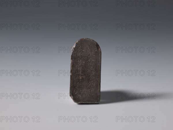 Stele-shaped tablet, Chinese, Qing dynasty 1644–1911, 19th century, China; stone slab with inscriptions; height 2.4 cm, width 5.15 cm, depth 0.75 cm; varia., A small upright rectangular object with a rounded top stands on a smooth surface, appearing dark brown to nearly black with a worn, slightly uneven texture and chipped edges; it casts a soft shadow to the right and is set against a smooth gradient background that shifts from darker gray at the top to lighter gray near the bottom.