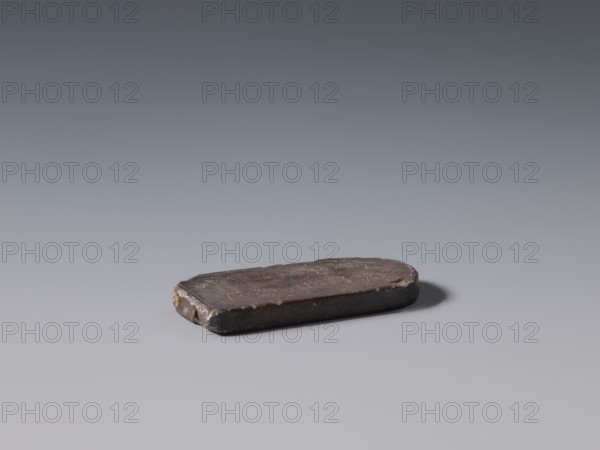 Stele-shaped tablet, Chinese, Qing 1644–1911, 19th century, China; stone slab with inscriptions; height 2.4 cm, width 5.15 cm, depth 0.75 cm., A small, flat, rounded rectangular object rests on a smooth surface, appearing dark brown with areas of lighter brown and hints of black; its top surface shows irregular texture and worn edges, and one end has a small patch of tan or ochre; the surface beneath casts a soft gray shadow and the background gradients from mid-gray near the object to darker gray toward the top.