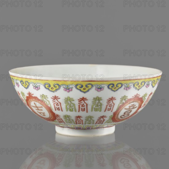 Bowl, porcelain on a ring foot; porcelain body with white glaze, decoration painted with overglaze enamel paints and iron red; colored porcelain; lambrequin pattern; longevity shou iconography; hieroglyphs; Qing dynasty, Jiaqing period early 19th century, 1796–1820; Chinese; height 5.8 cm, diameter 14 cm., A porcelain bowl with a white interior and exterior decorated with a band of yellow, blue, and pink ornamental motifs near the rim, a repeating pattern of red and green vertical decorative marks around the body, and larger circular cartouches in a reddish-orange hue spaced within the pattern; the bowl sits on a short white foot and casts a soft reflection on a smooth gray surface, with visible colors including white, yellow, blue, pink, red, green, reddish-orange, and gray.