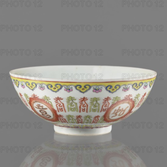 Bowl. Porcelain body with white glaze; decoration painted with overglaze enamel paints and iron red. Early 19th century Jiaqing period, 1796–1820, Qing dynasty 1644–1911, China. Colored porcelain bowl on a ring foot, height 5.8 cm, diameter 14 cm. Decoration: lambrequin pattern, longevity shou iconography, hieroglyphs; Chinese art/style., A shallow porcelain bowl sitting on a small foot with a reflective surface beneath, showing a mirrored underside; the exterior is predominantly white with decorative borders and patterns in yellow, blue, pink, green, and red, including a yellow rim band with blue and pink ornamental motifs and green and red vertical decorative elements around the body, plus circular red medallion-like designs; the interior appears pale white with subtle variations; the background is solid gray.