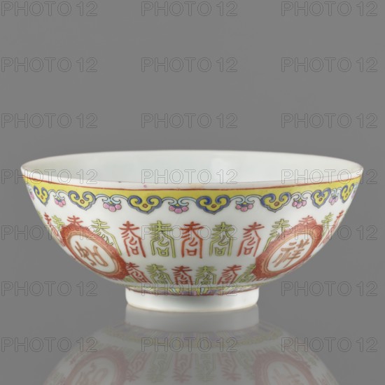 Bowl, Ceramic porcelain bowl on a ring foot with white glaze; decoration painted with overglaze enamel paints and iron red. Features lambrequin pattern, longevity shou iconography and hieroglyphs in a Jiaqing Jia-Qing Qing style. Colored porcelain, early 19th century Jiaqing period, 1796–1820, China. Height 5.8 cm; diameter 14 cm. ., A shallow porcelain bowl shown in profile against a gray background, with a glossy white interior and exterior decorated with painted motifs in colors including yellow, blue, pink, green, red, and orange; a decorative yellow and blue border runs near the rim accented with small pink elements, and below it a repeating vertical motif in green and red encircles the bowl interspersed with circular medallions outlined in orange containing a central decorative element; the bowl sits on a small foot and its underside is reflected faintly on the surface below.
