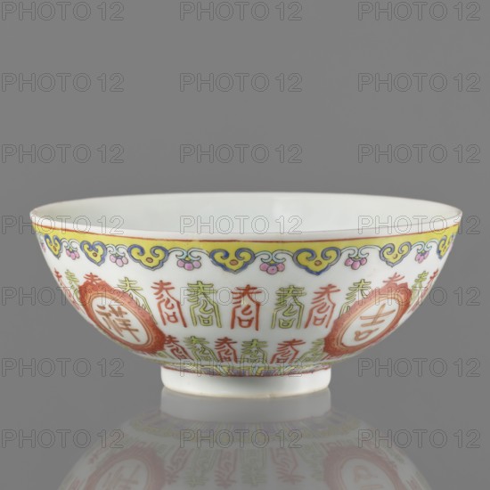 Bowl. Porcelain body; white glaze. Decoration painted with overglaze enamel paints and iron red, featuring longevity shou iconography, lambrequin pattern, and hieroglyphs. Colored porcelain, Qing dynasty, Jiaqing period early 19th century, 1796–1820. Bowl on a ring foot. Height 5.8 cm; diameter 14 cm., A white porcelain bowl with a rounded shape and a small foot sits against a neutral gray background, decorated around its exterior with a repeating painted pattern in red, green, yellow, blue, and pink, featuring ornamental motifs and circular medallions, and the interior rim showing a thin yellow border; the bowl casts a soft reflection on the surface beneath it.