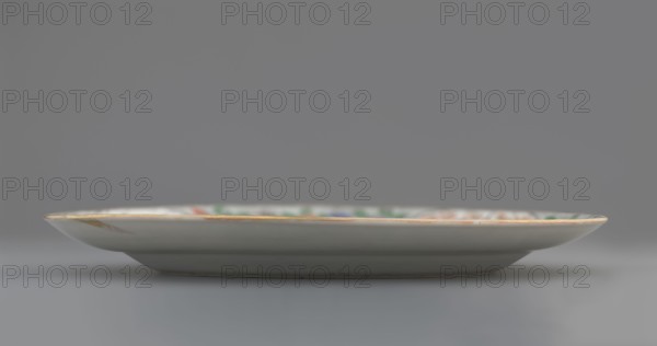 Plate. Porcelain fragment; white glaze. Decoration painted with overglaze enamel paints and iron red, colored porcelain with plant motifs and figural scenes. Qing dynasty 1644–1911, 18th century, China. Height 2.4 cm; diameter 20.4 cm; weight 0.35 kg., A shallow round ceramic plate shown from the side with a thin rim, a slightly raised foot, and a gently sloping profile; the plate surface appears mostly white or off-white with a narrow band of warm golden or tan color along the rim and hints of muted green, blue, and red visible near the inner edge; the background and surface beneath the plate are shades of gray.