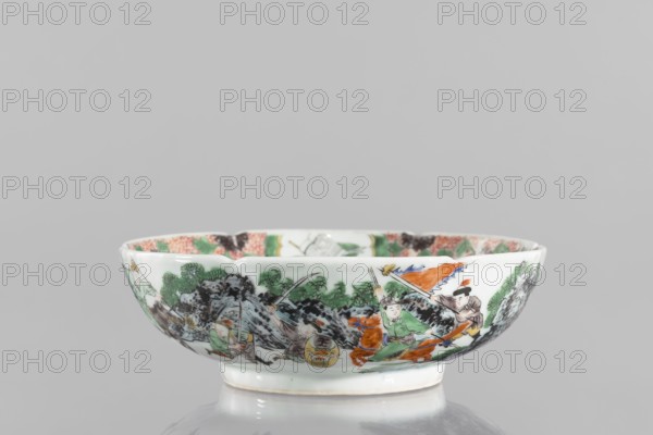 Bowl;, Porcelain body with white, transparent glaze; decoration painted with overglaze enamel paints and iron red. Chinese, Qing dynasty 1644–1911, 19th century. Colored porcelain with figural and knightly scenes; six-leafed belly. Vessel; height 6.5 cm, diameter 20.0 cm, weight 0.75 kg., A shallow porcelain bowl with a white base and a glossy finish sits against a neutral gray background, decorated around its exterior with a painted scene featuring a group of figures wearing green, orange, brown, and black garments, some holding long poles and a colorful banner, and accompanied by an orange horse; the scene includes stylized green foliage and dark rocky formations, with additional patterned decoration visible along the inner rim in shades of pink, green, and black.