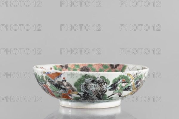 Bowl. Porcelain body with white, transparent glaze; decoration painted with overglaze enamel paints and iron red. Colored porcelain with figural and knightly scenes; six-leafed belly. Qing dynasty 1644–1911, 19th century, China. Height 6.5 cm; diameter 20.0 cm; weight 0.75 kg., A shallow porcelain bowl sits against a neutral gray background, showing a decorative painted scene around its exterior with a palette that includes green, black, orange, yellow, red, brown, and blue; the rim is white and the interior rim displays a patterned band in shades of green, red, and black; the exterior painting depicts a group of human figures wearing garments in green, orange, and brown tones, some holding long banners or staffs, positioned among stylized black and green foliage and rocky forms, with a glossy white foot at the base and a reflective surface beneath the bowl.