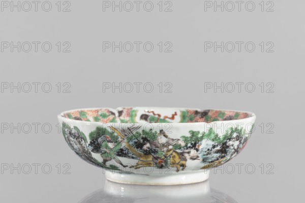 Bowl. Porcelain body with white, transparent glaze; decoration painted with overglaze enamel paints and iron red. Colored porcelain depicting figural scenes and knightly scenes; six-leafed belly. Qing dynasty 1644–1911, 19th century, China. Height 6.5 cm; diameter 20.0 cm; weight 0.75 kg.,  noted. Chinese., A shallow porcelain bowl with a white base color sits on a reflective surface, decorated around its exterior with a painted scene featuring a group of human figures and animals amid stylized landscape elements. The painted colors include green, black, brown, yellow, beige, pink, and hints of red and orange. The bowl has a slightly flared rim and a small foot, and the interior rim shows patterned decoration in similar colors. The surface appears glossy and smooth, and the background behind the bowl is a uniform light gray.
