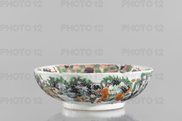 Bowl. Porcelain body with white, transparent glaze; decoration painted with overglaze enamel paints and iron red. Colored porcelain, figural and knightly scenes; six-leafed belly. Qing dynasty 1644–1911, 19th century, China. Height 6.5 cm; diameter 20.0 cm; weight 0.75 kg., A shallow porcelain bowl with a glossy white base is shown against a plain gray background; the exterior of the bowl is decorated with a painted scene in colors including green, orange, black, brown, and pale yellow, depicting human figures, foliage, and rocky forms in a continuous band around the rim, with green leafy clusters and black textured areas suggesting rocks or mountains, and orange elements that appear as garments and an animal figure, while the interior rim shows a patterned area with green and orange tones.