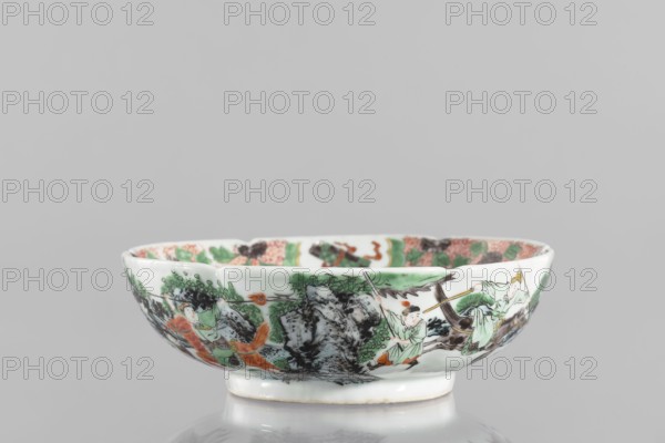 Bowl. Porcelain body with white, transparent glaze; decoration painted with overglaze enamel paints and iron red. Colored porcelain with figural scenes and knightly scenes; six-leafed belly. Qing dynasty 1644–1911, 19th century, China. Height 6.5 cm; diameter 20.0 cm; weight 0.75 kg., ; inscriptions/marks not specified., A shallow ceramic bowl with a glossy white base color, decorated around its exterior with a painted scene in green, black, orange, and hints of brown, depicting human figures in flowing robes amid stylized foliage and rocky formations; the rim shows a patterned interior band with green and orange elements and darker accents, and the bowl sits on a short circular foot with a slightly discolored edge.