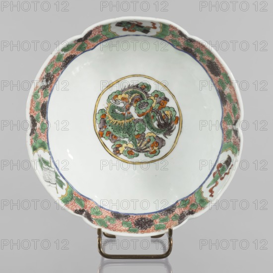 Bowl. Porcelain body with white, transparent glaze; decoration painted with overglaze enamel paints and iron red. Colored porcelain with figural and knightly scenes; six-leafed belly. Qing dynasty 1644–1911, 19th century, China. Height 6.5 cm; diameter 20.0 cm; weight 0.75 kg., A shallow round ceramic bowl with a scalloped rim sits on a small metal stand; the interior is mostly white with a central circular medallion containing a stylized creature rendered in green, orange, yellow, gray, and black surrounded by floral-like cloud motifs, and a narrow gold ring encircling the medallion; the rim interior is decorated with a patterned border featuring green leaves, coral red textured areas, and dark gray floral elements, with several small cartouche-like panels containing additional green, orange, and black motifs; the bowl shows subtle reflections and a smooth glazed surface against a neutral gray background.