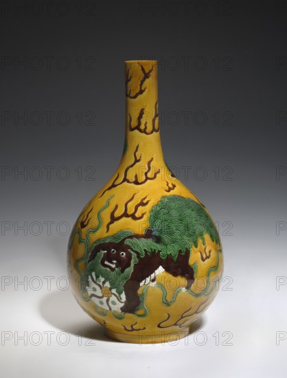 Onion-shaped vase, lion iconography, ceramics, Chinese art, Chinese culture, vases, Qianlong 1736–1795 style, pre-dated signatures. Vase with a narrow, long neck. Decoration on a yellow background: five lions—one larger with a ball and four smaller ones among flames. Yellow glaze, white in parts of the decoration; engraved decoration, painted with green and brown enamel paints. 19th century, China. Vessel. Height 32.0 cm; diameter 18.5 cm., A bulbous ceramic vessel with a long narrow neck sits against a dark gray background, the vessel painted a vivid yellow with flowing brown flame-like motifs; a stylized creature with a dark brown body and a textured green mane is depicted on the rounded body, standing over a pale circular object and surrounded by sinuous green tendrils, with highlights of white and small areas of lighter yellow on the base and shadowed white surface beneath.