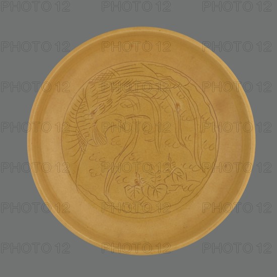 Plate, pheasants, ceramics, Chinese art, Chinese culture, Qing 1644–1911 style, monochromatic decoration, yellow; the mirror depicts a pheasant. Yellow glaze, finely crackled, engraved decoration, late 19th century, China, vessel; height 2.5 cm, diameter 14.6 cm., A round shallow dish with a smooth rim shown against a gray background, the dish is a warm mustard yellow with subtle darker speckling and a faint glazed sheen; inside the dish a lightly incised circular design depicts a stylized natural scene with flowing curved lines suggesting large leaves or fronds and rounded forms resembling a group of broad leaves or petals, with a few delicate linear marks indicating texture; the incised lines are slightly darker than the dish surface, and there are subtle variations of yellow and tan across the piece.