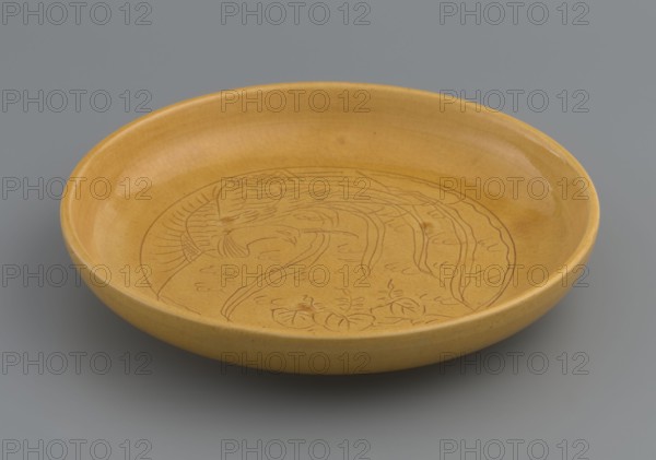 Plate, pheasants, ceramics, Chinese art, Chinese culture, Qing style, plates, Qing 1644–1911, Monochromatic decoration, yellow; the mirror depicts a pheasant. Yellow glaze, finely crackled, engraved decoration, late 19th century, China, vessel, height 2.5 cm, diameter 14.6 cm, A shallow round ceramic dish with a smooth glossy mustard-yellow surface sits against a gray background, showing a subtle darker yellow-brown line drawing of a stylized plant or floral motif inside the bowl; the rim and interior share the same yellow tone with slight variations and faint crackle patterns, and the underside edge is visible with a similar yellow color contrasted against the neutral gray surface beneath.