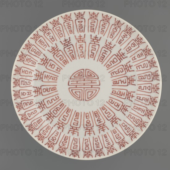 Plate;, Porcelain fragment with white glaze, decoration painted with overglaze iron red depicting longevity shou and bats. Colored porcelain, Qing dynasty, 19th century, China. Vessel; height 2.8 cm, diameter 18 cm, weight 0.2 kg., A round white ceramic plate sits against a gray background, decorated with a repeating radial pattern of red linear motifs arranged in concentric circles and a circular red emblem at the center; the design consists of several stylized geometric shapes and repeated vertical elements that radiate outward, creating a symmetrical, decorative effect in white, red, and gray.