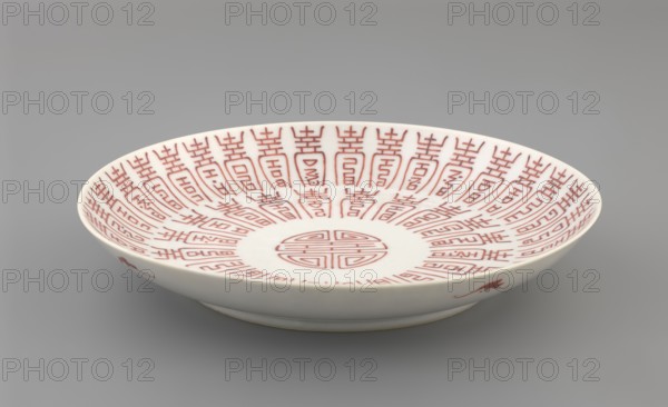 Plate., Porcelain fragment; white glaze with overglaze iron-red decoration of longevity shou character and bats. Colored porcelain, Qing dynasty, 19th century 1644–1911, China. Vessel. Height 2.8 cm; diameter 18 cm; weight 0.2 kg., A shallow white ceramic bowl with a wide rim sits on a smooth gray surface; the interior displays a repeated red linear pattern radiating from a central circular motif, and the bowl's exterior shows small red decorative marks near the rim.