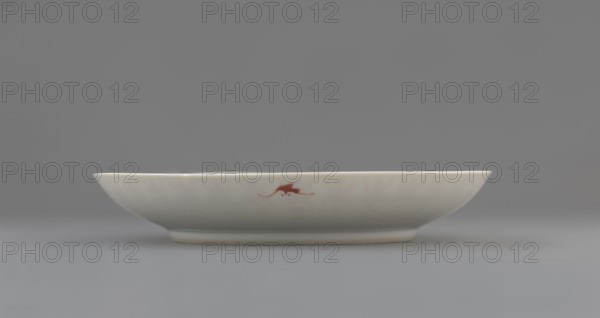Plate. Porcelain fragment with white glaze; decoration painted with overglaze iron red depicting longevity shou and bats. Colored porcelain, Qing dynasty, 19th century Qing, 1644–1911, Chinese. Vessel; height 2.8 cm, diameter 18 cm, weight 0.2 kg., function: plate., A shallow round ceramic dish sits on a flat surface, shown in profile against a neutral background. The dish is smooth and predominantly white with a glossy finish, and it has a small decorative motif in a reddish-brown color on its side. The background and surface are shades of gray, and the dish casts a soft shadow beneath it.
