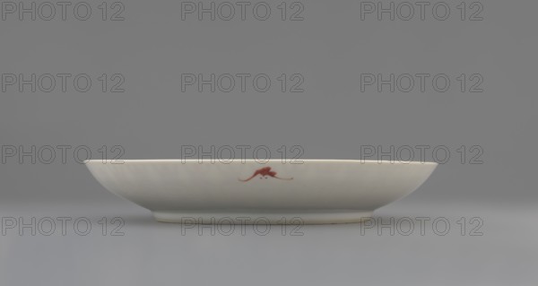 Plate;, Porcelain fragment with white glaze, decorated in overglaze iron red with longevity shou and bats iconography. Colored porcelain, Qing dynasty, 19th century, China. Vessel; height 2.8 cm, diameter 18 cm, weight 0.2 kg., A shallow white ceramic dish sits centered against a neutral gray background, showing a smooth curved rim and a slightly raised foot; the dish has a small reddish decorative mark on its side, and the visible colors in the image include white, gray, and reddish-brown.