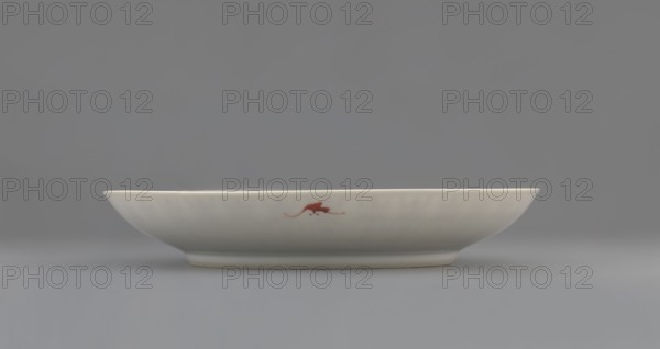 Plate;, Porcelain fragment with white glaze, decorated with overglaze iron red. Colored porcelain, Qing dynasty 19th century, China. Iconography: longevity shou and bats. Vessel; height 2.8 cm, diameter 18 cm, weight 0.2 kg., A shallow white ceramic dish with a smooth curved rim sits on a flat surface against a neutral background; the dish shows a small reddish-brown decorative mark near its center on the outer side and has subtle variations in white and off-white tones, while the surrounding background and surface appear in shades of gray and muted blue-gray.