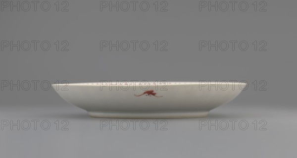 Plate, Chinese porcelain fragment with white glaze; decoration painted with overglaze iron red depicting longevity shou and bats iconography. Qing dynasty, 19th century 1644–1911. Vessel; height 2.8 cm, diameter 18 cm, weight 0.2 kg. Chinese art; colored porcelain; ceramics., A shallow round dish sits on a flat surface, shown in profile against a neutral background; the dish is primarily white with a smooth glossy finish and a subtle off-white or cream tone along its curved sides and base, and a small decorative brushstroke of reddish-brown on the visible side; the background and surface are shades of gray.