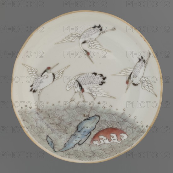 Plate. Chinese; Qing dynasty 1644–1911. Porcelain fragment with white glaze, decoration painted with overglaze enamel paints depicting cranes and waves. 19th century. Vessel. Height 2.8 cm; diameter 13.5 cm; weight 0.15 kg., A round ceramic plate with a pale cream background and a thin light brown rim, decorated with a scene of a group of white cranes with black feather accents and small red markings on their heads, depicted in various flying and standing poses. The lower portion shows stylized pale green and grey wave patterns, a bluish rock-like shape, and a rounded red form with white cloudlike details. The overall palette includes cream, white, black, red, pale green, grey, blue, and light brown.