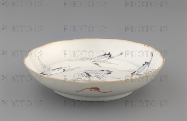 Plate. Chinese; Qing dynasty 1644–1911. Porcelain fragment with white glaze and overglaze enamel-painted decoration depicting cranes and waves. 19th century. Vessel; height 2.8 cm, diameter 13.5 cm, weight 0.15 kg., A shallow white ceramic bowl with a slightly scalloped rim rimmed in a warm golden-beige color, decorated inside with a group of stylized birds rendered in black and gray line work with small areas of muted red on their heads and beaks, and a subtle red ornamental motif on the exterior side; the interior glaze appears glossy white with delicate fine brush detailing on the birds and faint beige discoloration at the rim, set against a neutral gray background.