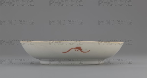 Plate. Chinese porcelain fragment, Qing dynasty 1644–1911, 19th century. White glaze with overglaze enamel-painted decoration depicting cranes and waves. Vessel fragment; height 2.8 cm, diameter 13.5 cm, weight 0.15 kg. Maker/craftsman unknown., A shallow ceramic dish sits horizontally against a neutral background, showing a smooth white surface with a subtle glossy finish and a scalloped rim accented with a thin golden edge. A small decorative flourish in reddish-brown appears on the side of the dish, consisting of a curved line with a rounded central element. The dish rests on a short circular foot, and the visible colors in the image include white, gray, gold, reddish-brown, and muted bluish-gray tones in the background.