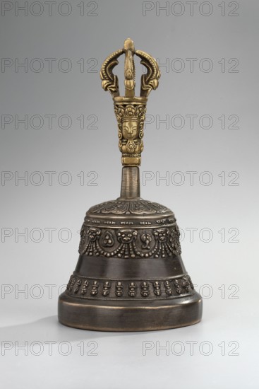 Bell, Tibetan Buddhist art, Himalayan art, Mongolian art, Himalayan culture, Mongolian culture, vajras, musical instruments, percussion instruments, idiophones, religious rites, ritual object, late 18th century, Mongolia place of origin? — ceremonial bell, height 15.7 cm, diameter 8.2 cm, A decorative handbell sits upright on a neutral gray surface against a pale gray background, with a metallic appearance combining dark bronze and golden tones; the bell body is dark bronze with ornate raised patterns and beaded borders encircling it, and the handle is golden with intricate carved faces and curved ornamental shapes at the top.