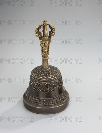 Bell, Tibetan Buddhist art, Himalayan art, Mongolian art, Himalayan culture, Mongolian culture, vajra, musical instrument, percussion instrument, idiophone, used in religious rites, ritual object, late 18th century, Mongolia place of origin ?, ceremonial object; height 15.7 cm, diameter 8.2 cm, A decorative metal bell sits on a smooth pale gray surface, featuring a dark bronze body with ornate raised patterns and relief bands, and a vertical handle topped by a golden ornate finial; the bell shows textured floral and beaded motifs on its rounded surface, a narrower ribbed section leading to the handle, and a shadow on the surface beneath it.