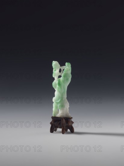 Guanyin, unknown creator; white-green jade; full sculpture, engraved; figurine of a goddess from Chinese Buddhism; Qing dynasty, 19th century, China; height 6.0 cm., A vertical carved object with smooth, flowing contours sits on a small dark wooden stand; the carved piece shows a blend of pale green and white tones with rounded, bulbous forms and some open spaces within its shape, while the stand is brown with a slightly irregular, organic silhouette and short legs, all set against a gradient background that shifts from dark at the top to light gray at the bottom.