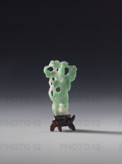 Guanyin, white-green jade, full sculpture, engraved, 19th century Qing dynasty, China, Chinese Buddhist goddess, fan, figurine, height 6.0 cm., A carved object stands on a small stand against a dark to light gray gradient background; the carved piece is a translucent pale green with areas of lighter and darker green and some white, shaped with organic flowing forms and several rounded openings, and the stand is a dark brown with a slightly irregular, sculpted shape.