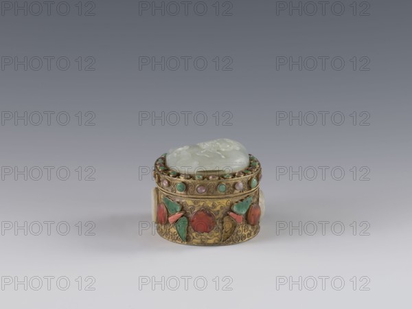 Box with lid, Chinese, Qing dynasty late 19th century. Lid inlaid with a jadeite plaque with a bas-relief image of a fo lion playing with a ball. Outer wall decorated with motifs of butterflies, bats, and the shou longevity symbol made of semi-precious stones set in settings. Background: plant twig with filigree. Vessel/container; height 5.0 cm, width 6.6 cm, depth 8.2 cm, weight 0.17 kg., A small round decorative box sits on a smooth surface against a gradient gray background; the box has a metallic gold base with ornate engraved patterns and is set with several circular stones in red, green, and pink hues and small turquoise accents, with a pale translucent lid that appears white to light green and shows carved floral-like shapes.