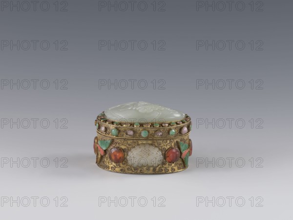 Box with lid, Chinese art, Chinese, Qing 1644–1911. The lid is inlaid with a jadeite plaque with a bas-relief image of a fo lion playing with a ball. The outer wall features motifs of butterflies, bats, and the shou symbol of longevity made of semi-precious stones set in settings. Background: plant twig with filigree. Late 19th century, China. Vessel/container. Height 5.0 cm, width 6.6 cm, depth 8.2 cm, weight 0.17 kg., A small ornate oval box sits centered on a gradient gray background, with a pale green carved lid and a gold-toned body decorated with a few round cabochon stones in green and reddish-orange, several small green and pink circular insets along the upper rim, and carved pale panels set into the sides; the gold surface shows intricate raised scrollwork and floral motifs, and there are a few green and coral-colored leaf-shaped accents on the sides.