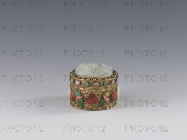 Box with lid;, Chinese, Qing dynasty 1644–1911, late 19th century. The lid is inlaid with a jadeite plaque with a bas-relief image of a fo lion playing with a ball. The outer wall features motifs of butterflies, bats, and the shou symbol of longevity made of semi-precious stones set in settings. Background: plant twig with filigree. Vessel/container. Height 5.0 cm; width 6.6 cm; depth 8.2 cm; weight 0.17 kg., A small round decorative box sits centered on a smooth gradient background, with a pale green carved lid resting on an ornate golden body; the gold surface is decorated with a pattern of swirls and set with several small round stones in shades of green and pink and a few larger rounded cabochons in red and coral tones, along with green leaf-shaped inlays and pale cream circular side accents.
