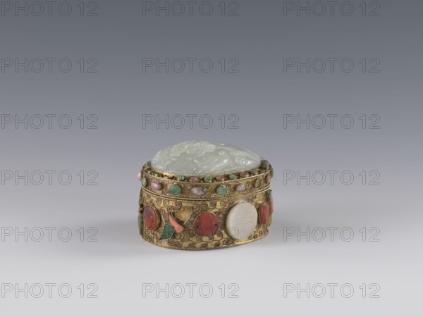 Box with lid, Chinese, Qing dynasty late 19th century. Height 5.0 cm, width 6.6 cm, depth 8.2 cm, weight 0.17 kg. The lid is inlaid with a jadeite plaque with a bas-relief image of a fo lion playing with a ball. The outer wall features motifs of butterflies, bats, and the shou symbol of longevity made of semi-precious stones set in settings. Background: plant twig with filigree. Vessel/container., A small oval decorative box sits on a smooth surface against a gradient gray background, with a domed pale green lid carved with raised motifs and a body of ornate gold metalwork embellished with a group of round and oval cabochon stones in colors including green, red, pink, white, and turquoise, along with small green and pink bead accents and filigree scrollwork in the gold.