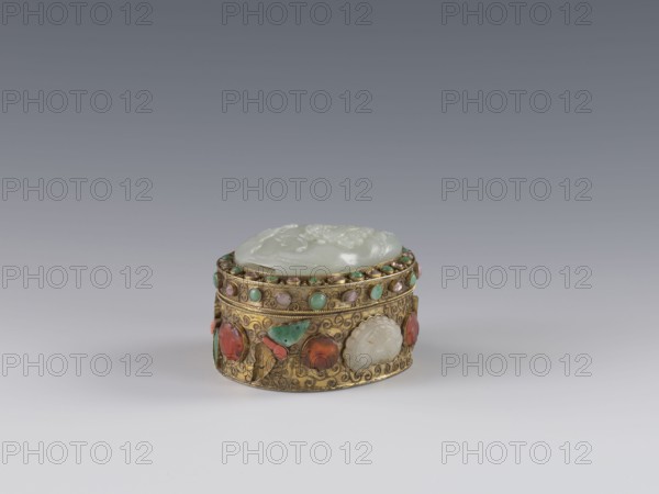 Box with lid;, Chinese, Qing dynasty 1644–1911, late 19th century. Jadeite plaque inlaid in the lid with a bas-relief image of a fo lion playing with a ball. Outer wall decorated with motifs of butterflies, bats, and the shou symbol of longevity made of semi-precious stones set in settings; background shows a plant twig with filigree. Function: vessel/container. Materials: jadeite and semi-precious stones. Dimensions: height 5.0 cm, width 6.6 cm, depth 8.2 cm, weight 0.17 kg., A small ornate circular box sits centered on a smooth surface, featuring a pale translucent lid with carved raised details and a patterned gold-colored band around the sides decorated with a few rounded green, pink, and red stones and several carved pale insets; the metal band shows intricate scrolling designs and small beaded accents. The scene includes a gradient background shifting from mid-gray at the top to light gray near the surface, and the predominant colors visible are gold, pale white, green, pink, red...