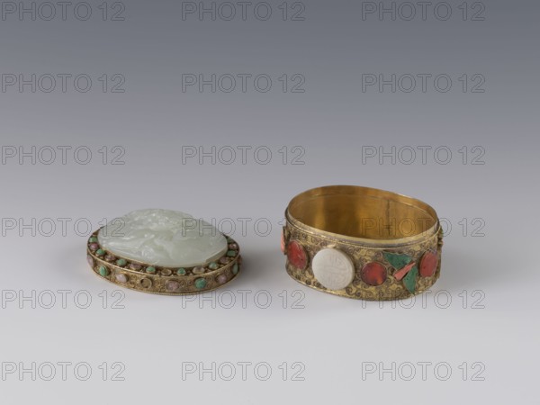 Box with lid, Chinese, Qing 1644–1911, late 19th century. The lid is inlaid with a jadeite plaque with a bas-relief image of a fo lion playing with a ball. The outer wall features motifs of butterflies, bats, and the shou symbol of longevity made of semi-precious stones set in settings. Background: plant twig with filigree. Vessel/container; height 5.0 cm, width 6.6 cm, depth 8.2 cm, weight 0.17 kg., A small decorative box with a separate lid sits on a neutral surface; the lid is oval, pale green and carved with a raised floral or foliate motif, and its rim is set with a band of small round stones in green, pink, and beige hues set in a gold-colored metal frame; beside it is a matching oval container in a gold-colored metal with an open interior showing a warm golden tone, and the outer band is decorated with several cabochon stones in red, green, and pale green colors as well as carved pale green oval elements, all set against a textured gold surface with scrolling patterns; the backg...