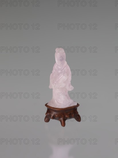 Guanyin on a base, figurine carved in rose quartz, full sculpture with engraving; goddess of Chinese Buddhism; Chinese art, Qing dynasty 1644–1911; 19th century; sculptor, height 6 cm., A small translucent pink figurine of a robed person standing on a low brown wooden stand, with the pink surface showing smooth carved folds and facial features, set against a uniform gray background with a faint reflection below; visible colors include pink, brown, and gray.
