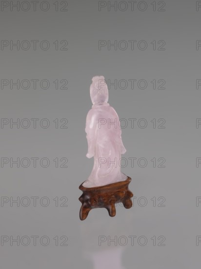 Guanyin on a base, Chinese art, Chinese culture, Qing 1644–1911, Chinese Buddhism, goddess, figurine carved in rose quartz, full sculpture, engraving, 19th century, China, sculpture, height 6 cm, A translucent pink carved figure standing on a small wooden stand with a polished brown finish; the figure appears to wear draped clothing and has a textured hairstyle, and the background is a smooth gradient of gray with reflections of the pink and brown tones visible on the surface below.