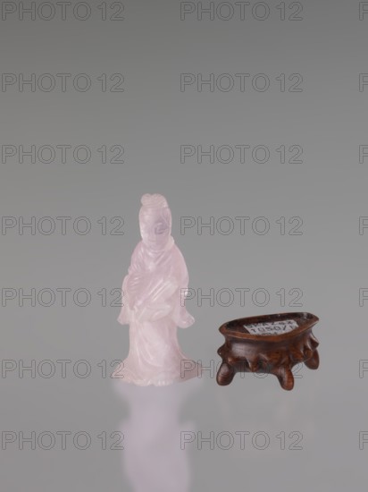 Guanyin on a base; figurine carved in rose quartz; full sculpture with engraving; Chinese; Qing dynasty 1644–1911; Chinese Buddhism; goddess; 19th century; sculptor, height 6 cm., A small translucent pink figurine of a robed person standing beside a low dark brown wooden stand with short rounded legs; the background is a smooth gradient of gray and the surface beneath reflects faintly with soft pink and gray tones.