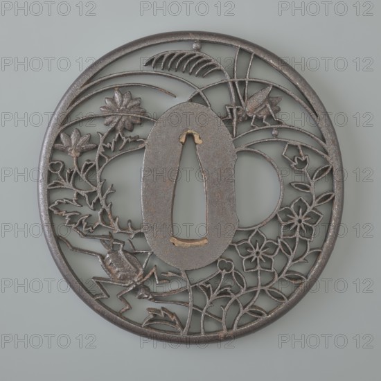 Crossguard tsuba, Akasaka role classification of creator, school author; Japanese; Edo period 1603–1868, 18th/19th century. Tsuba round maru gata with a rounded edge maru mimi; seppa dai oval; sekigane copper. Decorated with openwork ji sukashi decoration, partly positive and partly negative, depicting two crickets among chrysanthemums, flowers, and grasses. Not signed. Function: sword crossguard katana. Diameter 7.2 cm; thickness 0.5 cm., A round metal object with a dark gray to brownish patina sits against a pale gray background; the piece has an openwork design featuring a central oval cutout framed by a ring of thin metal, with floral motifs and leafy vines woven throughout the circular field, a few stylized flowers and several curved leaf shapes, and a couple of insect-like forms rendered in the same dark metallic tones.