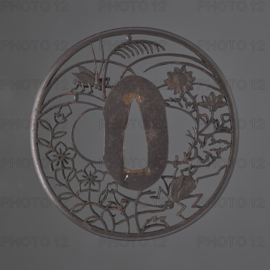 Crossguard tsuba, Akasaka role classification of creator, school author. Japanese, Edo period 18th/19th century. Tsuba round maru gata with a rounded edge maru mimi; seppa dai oval; sekigane copper. Decorated with an openwork ji sukashi design, partly positive and partly negative, depicting two crickets among chrysanthemums, flowers, and grasses. Not signed. Diameter 7.2 cm; thickness 0.5 cm., A round metal object with an openwork floral and insect design set against a gray background; the object is dark brown to black with areas of rust-colored brown and small patches of lighter brown. The design includes a central oval opening surrounded by cut metal stems, leaves, and several stylized flowers, and a few insect shapes incorporated into the pattern. The outer rim is circular and the metal has a textured, aged surface with subtle variations of dark gray and brown.