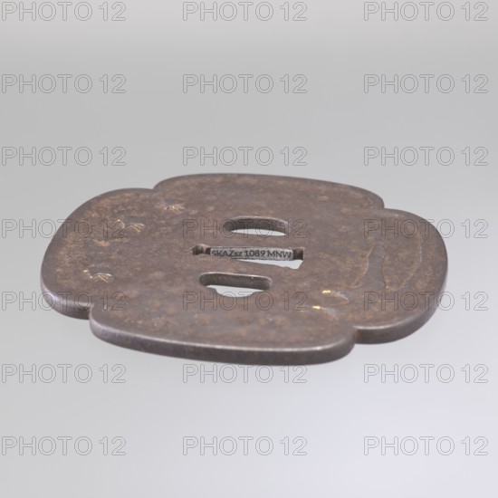 Crossguard tsuba, Kaneie; geese; sword crossguards; mokkô; Japanese art; Japanese culture; swords; katana; weapons; bladed weapons; Edo period 1603–1868; birds; grasses; boats. Tsuba for a tachi sword. Quatrefoil shape mokkô gata with a slightly raised edge of the uchi kaeshi / ryôhitsu ana type. Decorated on the obverse with wild geese flying over a water's edge overgrown with reeds; on the reverse, sails of three boats at the top and fishing nets drying on the shore below. Gold-inlaid details. Signed. Early 19th century Edo period. Fushimi, Yamashiro province, Japan. Tsuba; height 10.9 cm, width 10.2 cm, depth 0.45 cm., A flat, rounded metallic object with an irregular flower-like outline rests against a neutral gray background; the metal surface is dark brown with mottled rust tones and small patches of lighter brown and faint golden flecks, and it has a few rounded cutouts and an elongated horizontal opening near the center, along with subtle raised and recessed areas indicating sh...