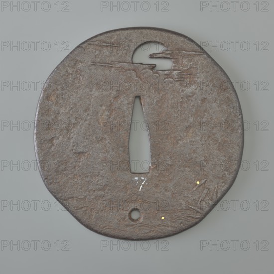Crossguard tsuba, Kaneie,  school craftsman author. Oval tsuba with an irregular outline slightly merging into the shape of a quatrefoil mokkô, slightly marked edge uchi kaeshi, textured surface. Decorated in low relief with details inlaid with gold and cut into negative openwork. On both sides a similar motif of waterside reeds and a full moon emerging from behind the clouds. At the bottom, one hole udenuki ana. Signed. 18th century Edo period, 1603–1868. Japan; Fushimi, Yamashiro. Materials/subject: Japanese, moon iconography, weapons/melee weapons/swords/katana/sword crossguards/tsuba. Dimensions: height 7.7 cm; width 7.6 cm; depth 0.3 cm., A round, flat metal object with a weathered, rough surface in brown and rusty tones, featuring a central elongated vertical aperture and a few circular cutouts; the metal shows irregular texture and patina with small areas of darker brown and lighter grey, and there are a few tiny bright yellowish spots on the lower right area, along with subtle ...