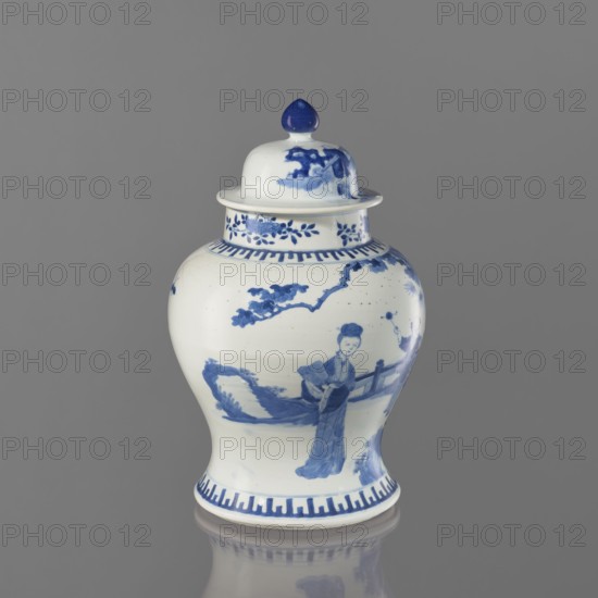 Vase with lid, Chinese, 19th century, thick porcelain body, greyish-blue glaze with underglaze cobalt painted decoration; decorated with boys, butterflies, female figures, and terraces architectural motifs; vessel, height 36.5 cm., A white ceramic jar with a lid featuring blue painted decorations sits against a gray background, showing a glossy surface and a reflection beneath it; the jar has a rounded body that tapers at the base and neck, a domed lid topped with a rounded finial, and blue decorative bands near the rim and base; the body displays blue-painted scenes including a standing figure in traditional clothing, branches with leaves, and landscape elements rendered in blue tones.